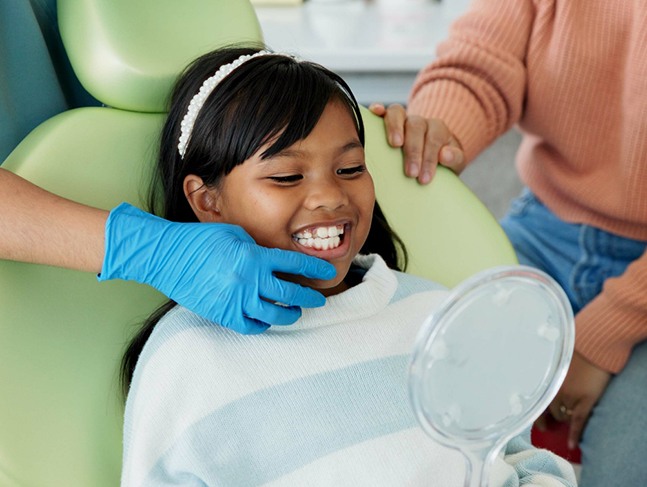 Dentist touching little girl’s chin as she looks at her teeth in a handheld mirror